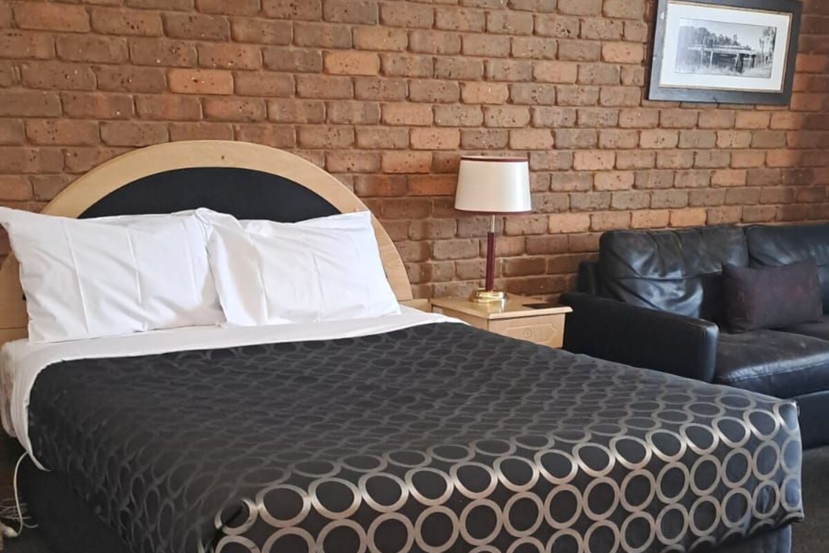 Una habitación de hotel con pared de ladrillo, una cama con colcha estampada, una mesita de noche con lámpara y un sofá de cuero.