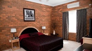 A bedroom with a brick wall, a bed with a dark headboard, two bedside tables with lamps, a window with curtains, and a flat-screen TV.