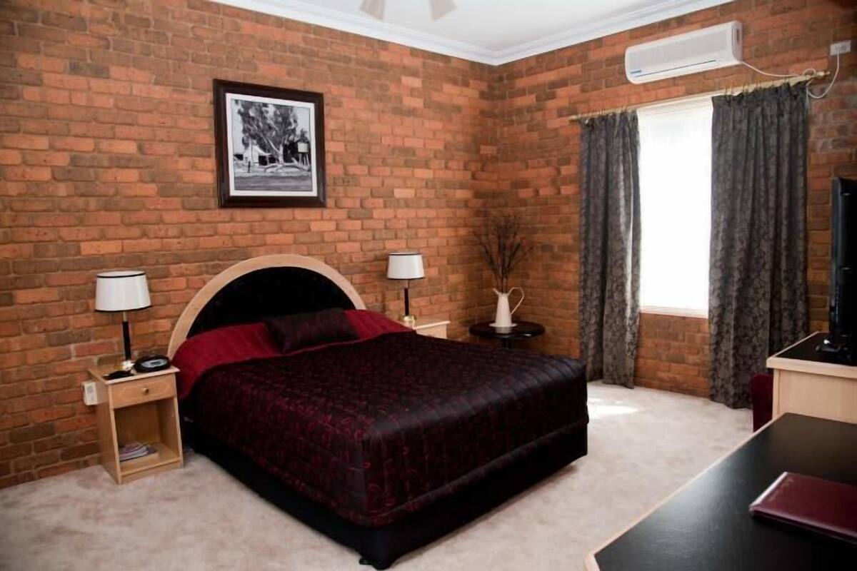 A bedroom with a brick wall, a bed with a dark headboard, two bedside tables with lamps, a window with curtains, and a flat-screen TV.