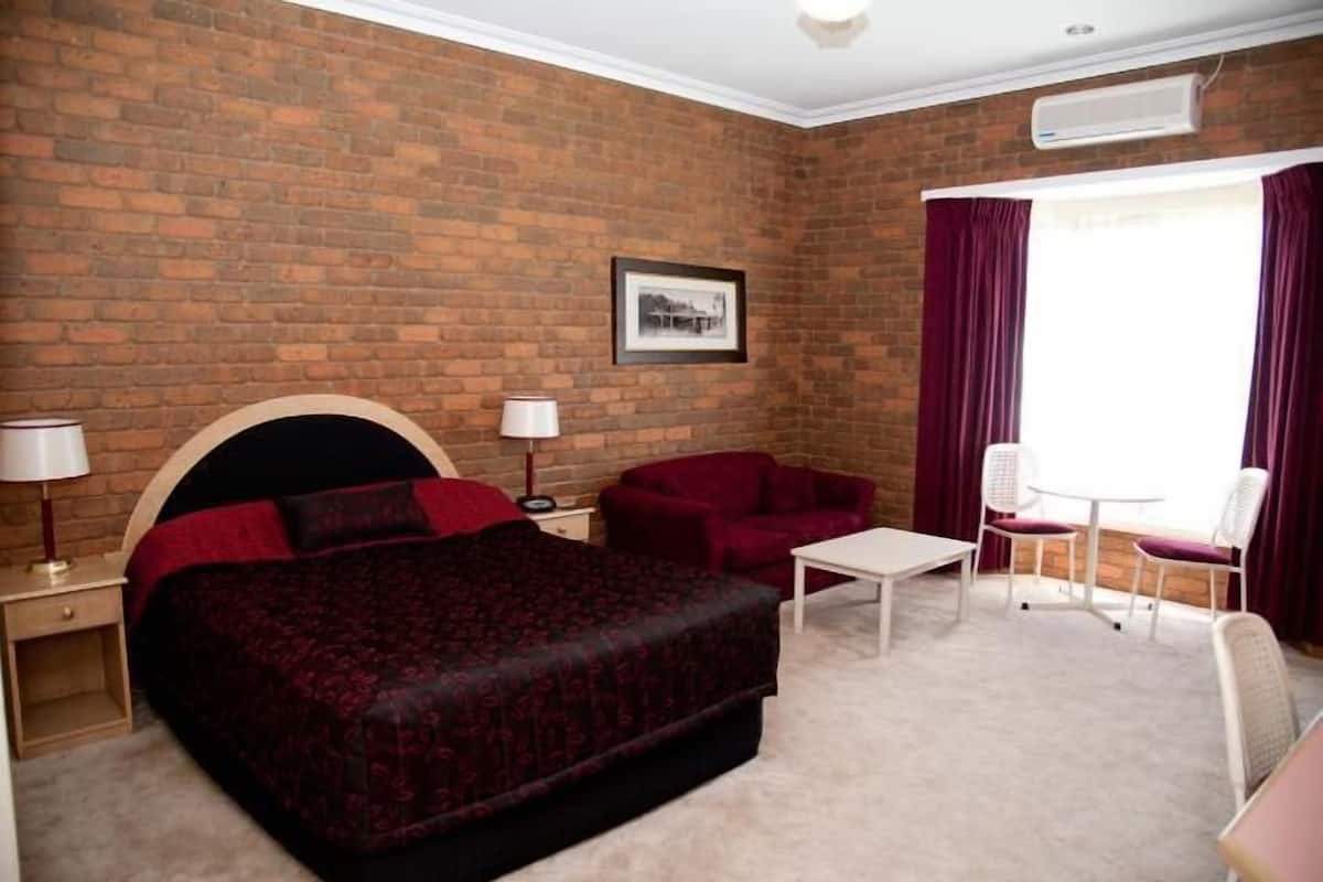 A hotel room with a brick wall, a bed with a headboard, a small table, two chairs, and a window with curtains.