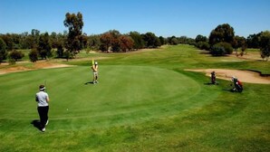 A golf course with a clear sky, a golfer preparing to hit a ball, and a caddy with a golf bag.