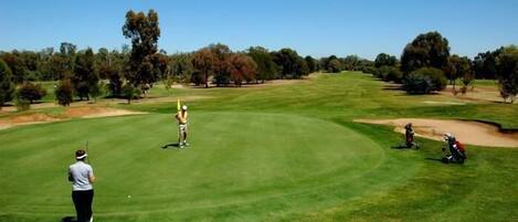 Un campo de golf con cielo despejado, un jugador de golf preparándose para golpear la bola, y un caddie con un bolso de golf.