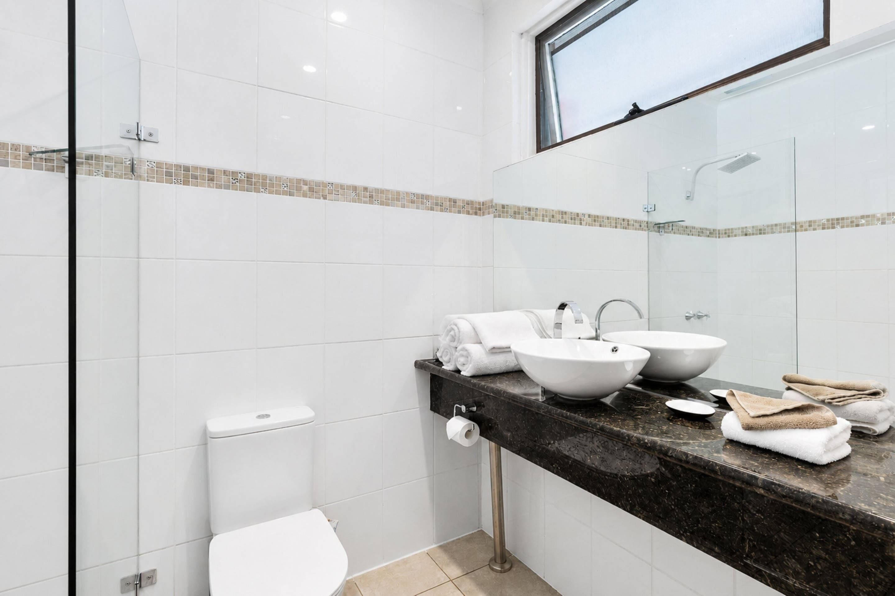 Ein modernes Badezimmer mit weißen Fliesen, einer Glasdusche und einer Marmorarbeitsplatte mit zwei Waschbecken und Handtüchern.