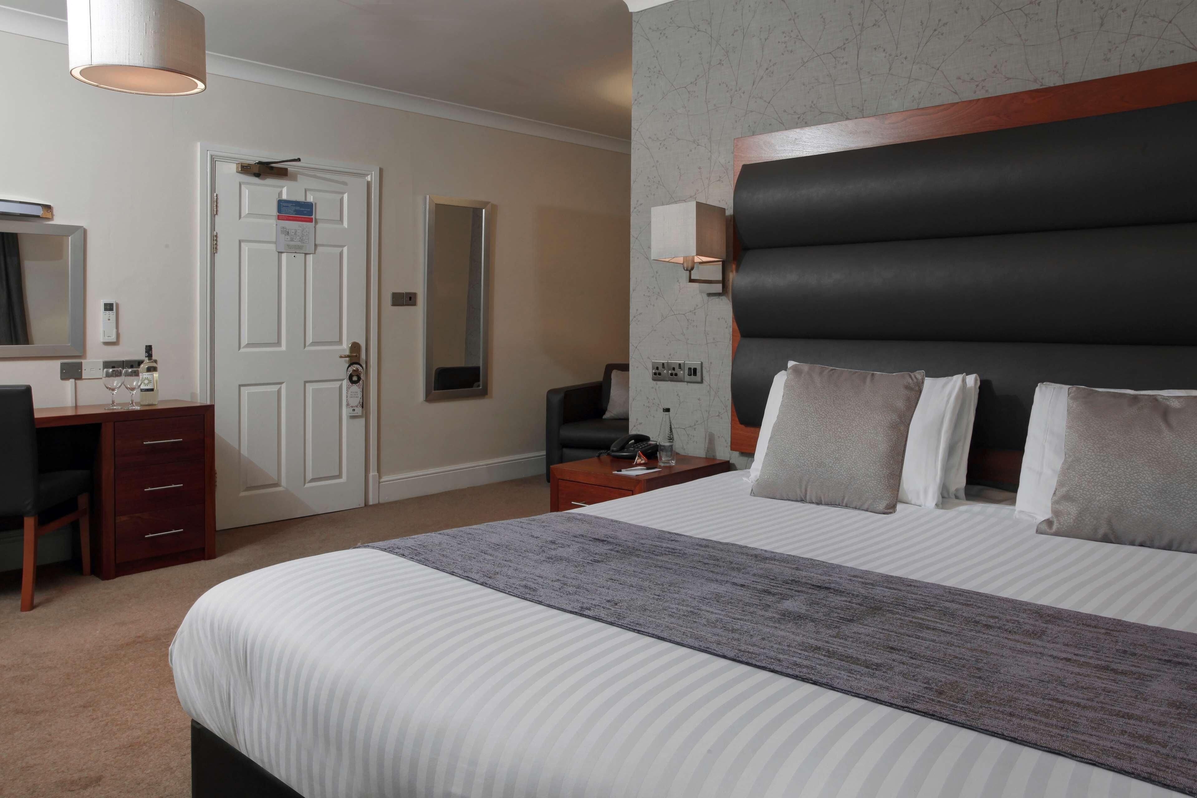 A hotel room with a bed, a desk, a chair, a dresser, a nightstand, a lamp, a mirror, and a door.