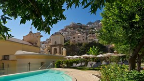 Zona de piscina con sillas de descanso y sombrillas, una escalera que lleva a un edificio y vistas a una colina con construcciones.