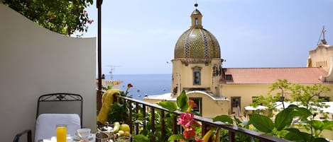 Un balcón con una mesa preparada para el desayuno, con vistas a la costa y una iglesia con cúpula.