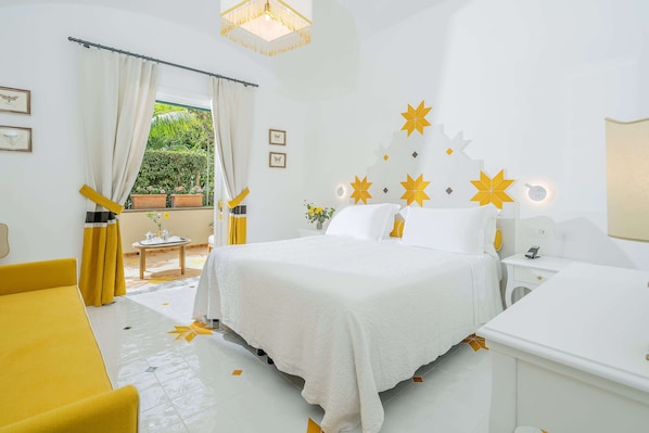A hotel room with a bed, a bedside table, a chair, a window with curtains, and a view of greenery outside.