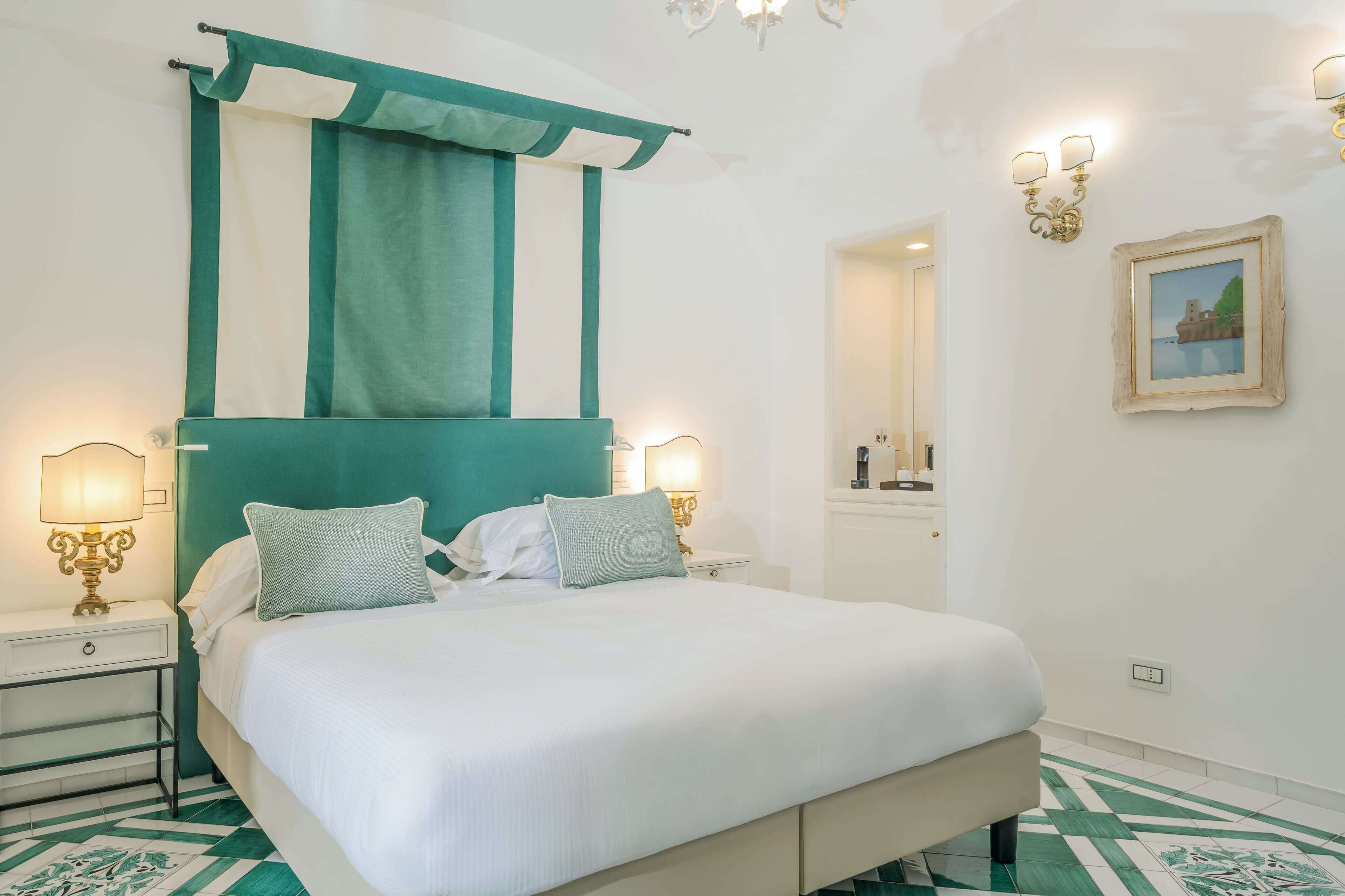 A neatly made bed with white linens and two pillows, a teal accent headboard, and a nightstand with a lamp.