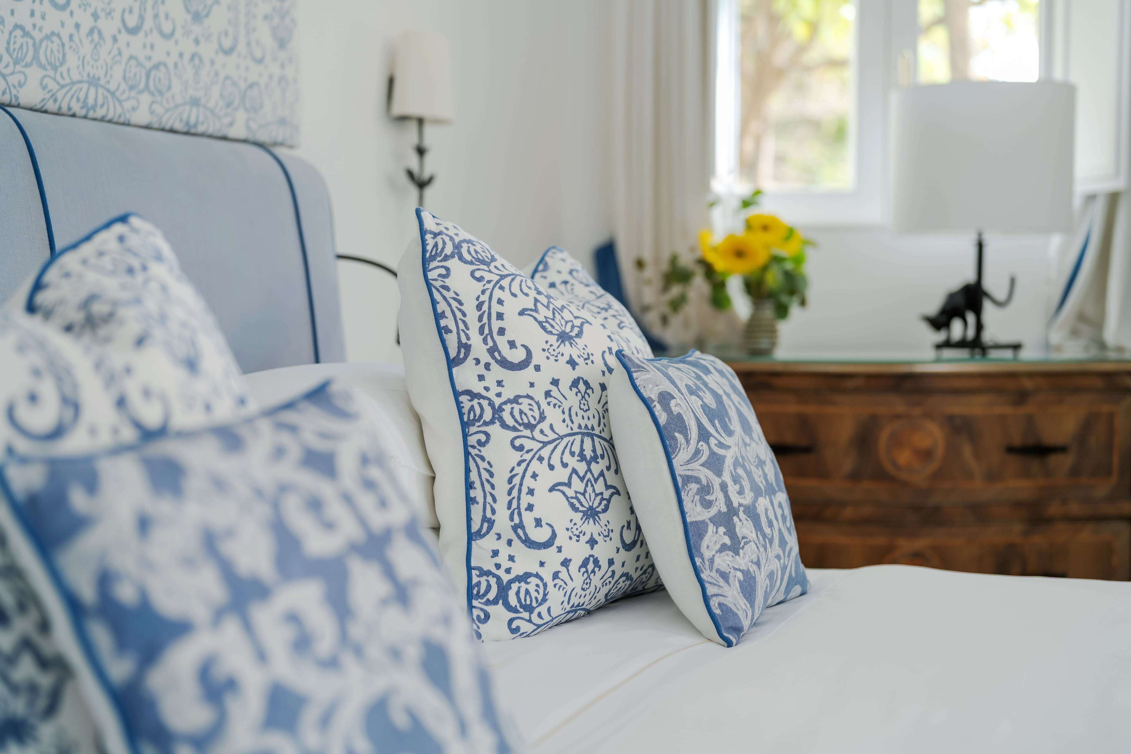 A bedroom with a bed, blue and white patterned pillows, a wooden dresser, a vase with flowers, and a window with curtains.