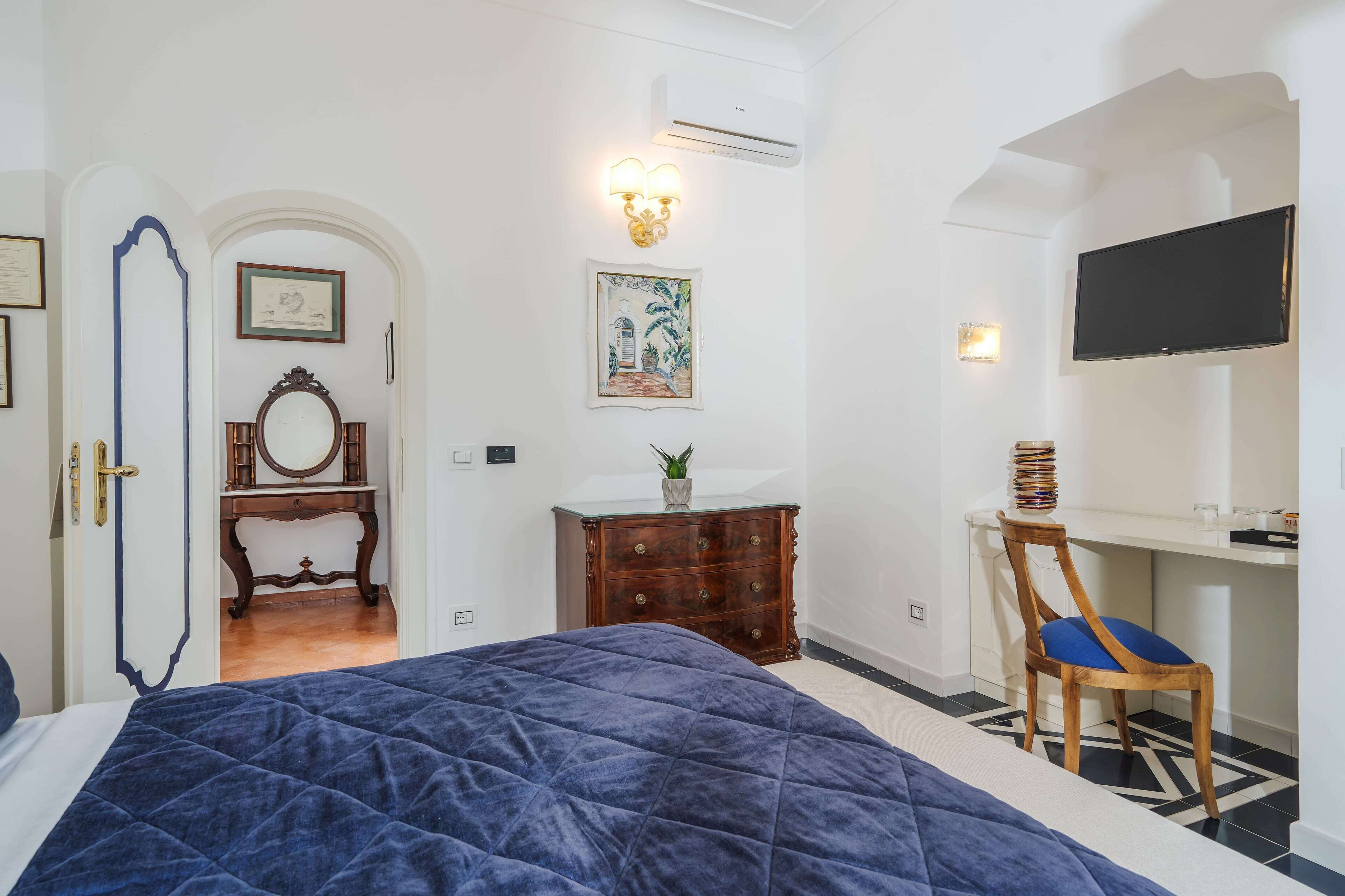 Un dormitorio con una cama grande, un aparador de madera, un escritorio con una silla, un televisor montado en la pared y una planta pequeña sobre el aparador.