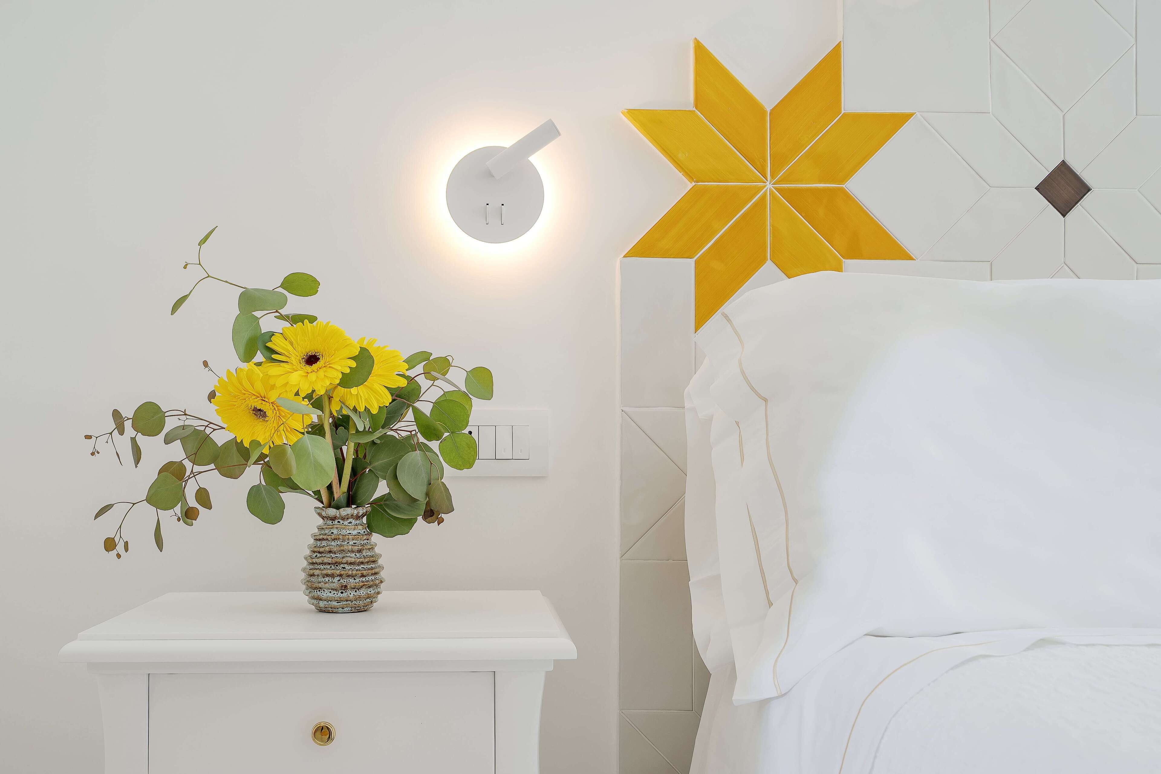 A white bedside table with a vase of yellow flowers, a wall-mounted light, and a geometric wall decoration.