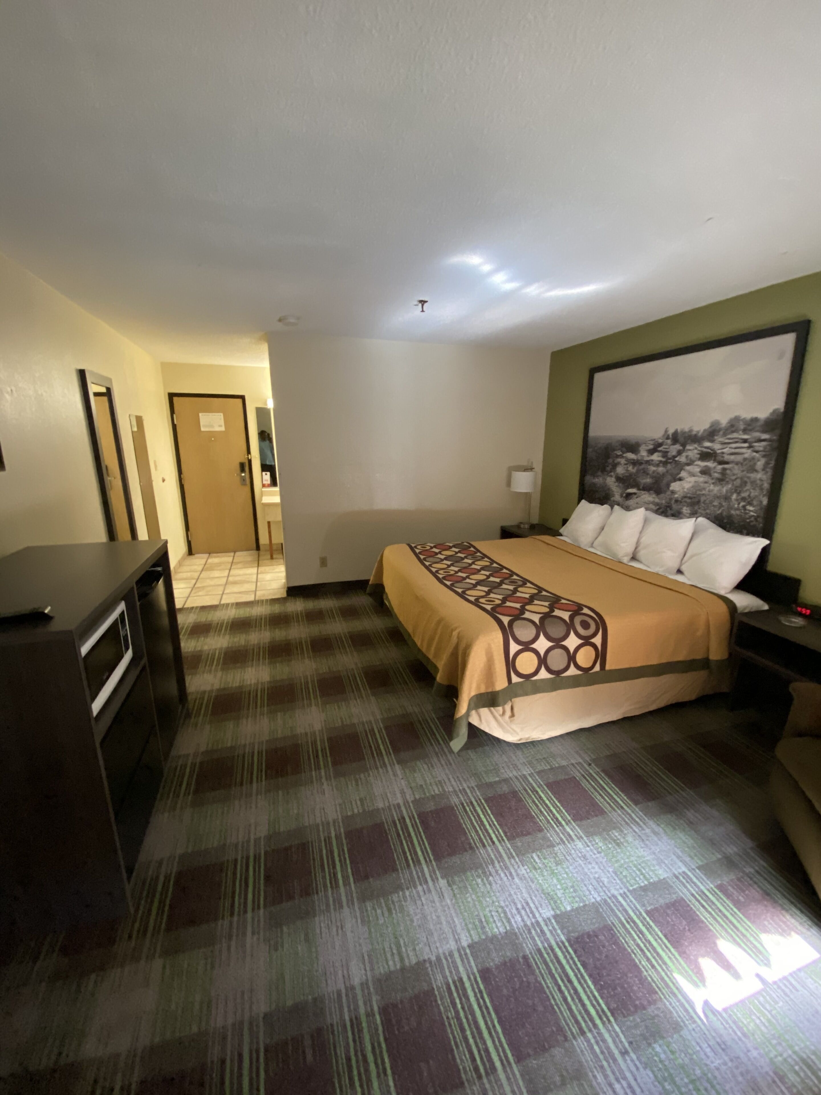 Habitación de hotel con cama, escritorio, silla, mesita de noche, lámpara y un cuadro en la pared.