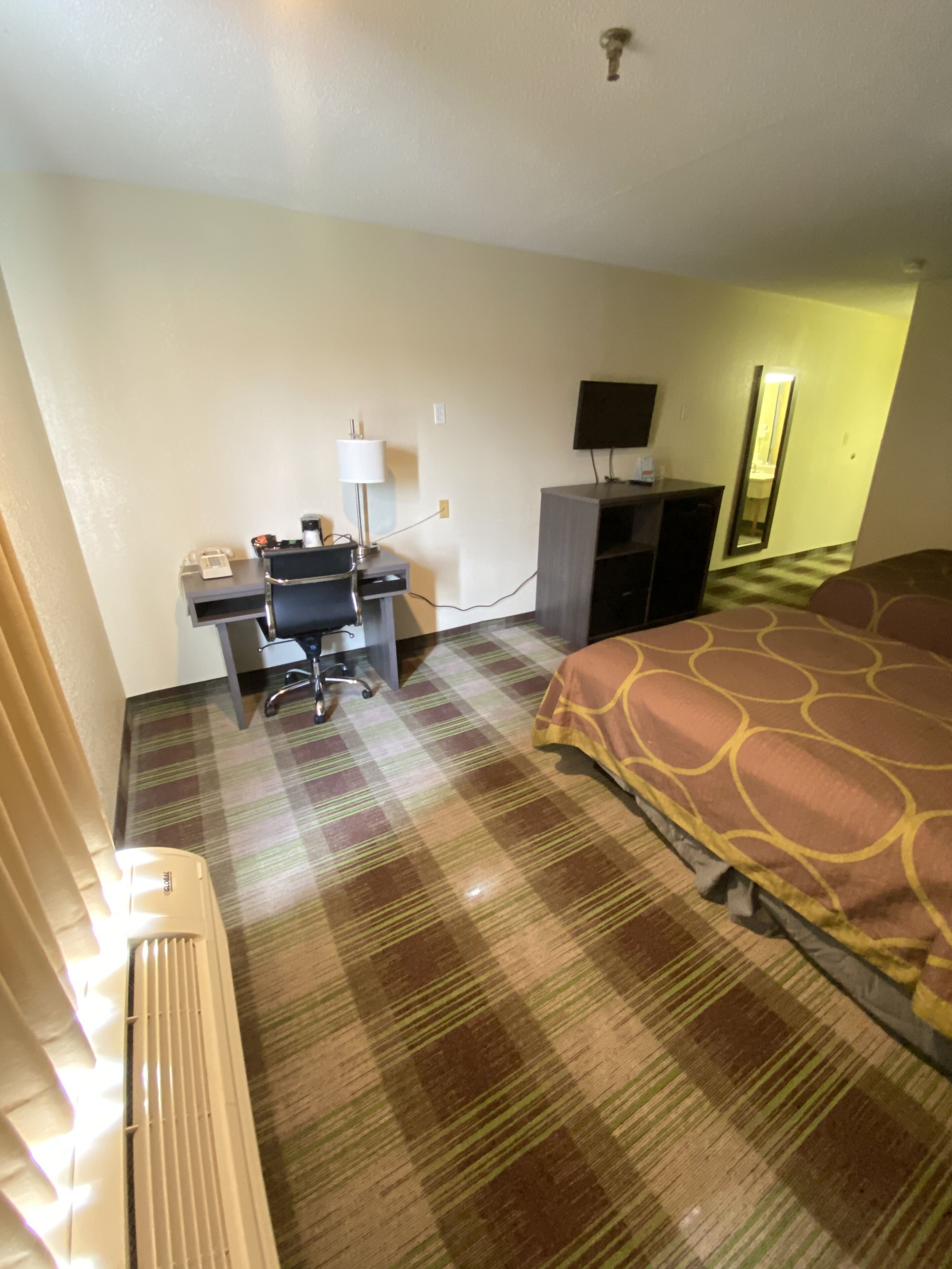 Una habitación de hotel con cama, escritorio con computadora, silla, lámpara, televisor y espejo.