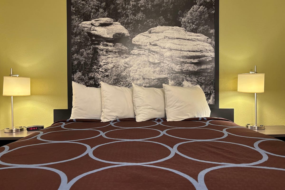 A hotel room with a bed, two bedside lamps, a digital clock, and a large rock wall painting.