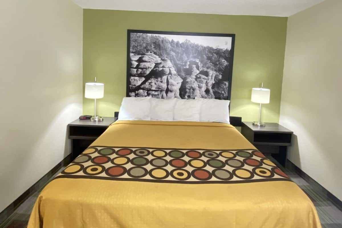 A hotel room with a bed, two bedside tables with lamps, a wall-mounted light fixture, and a framed picture above the bed.