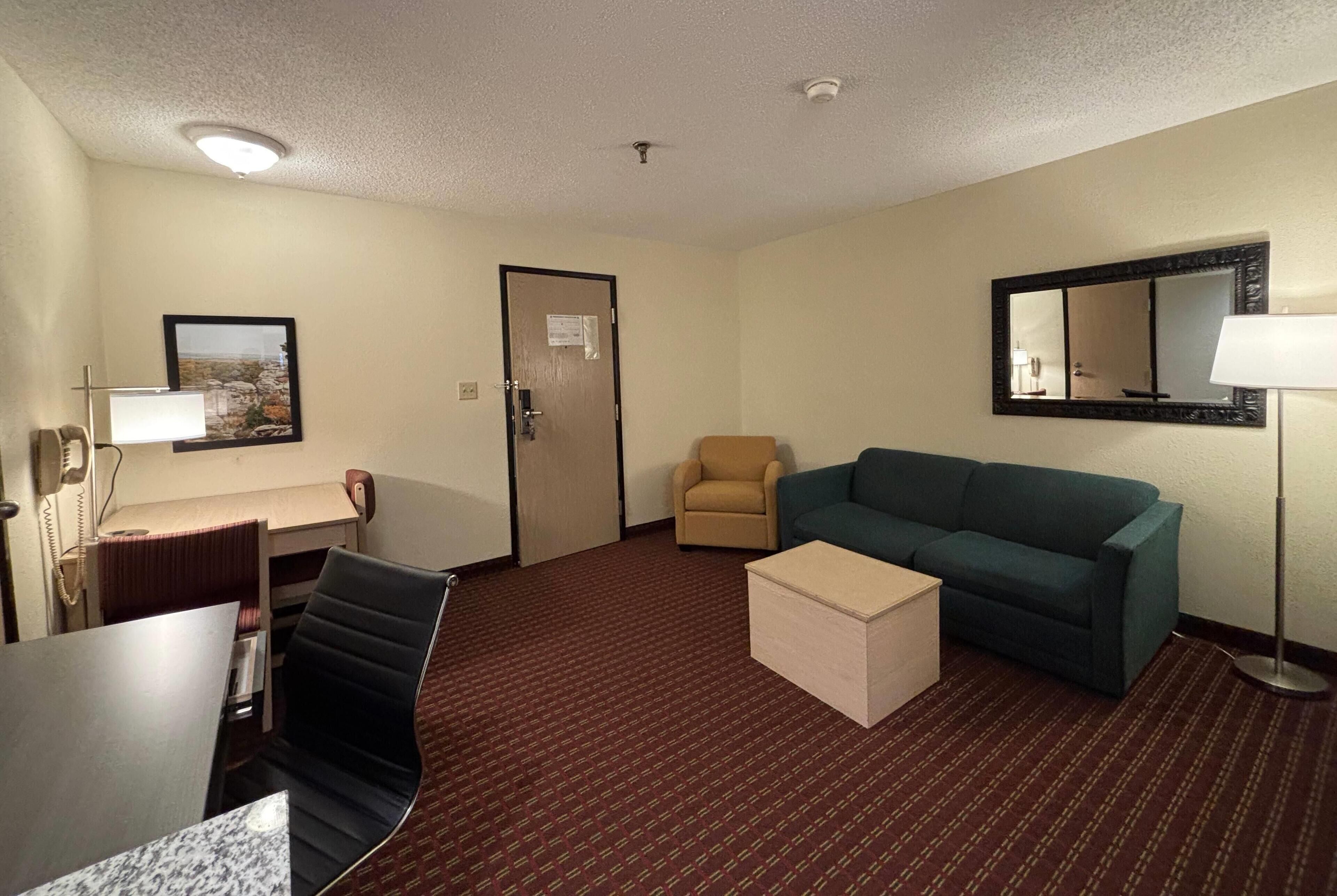 A hotel room with a desk, a chair, a sofa, a small table, a lamp, a mirror, and a door.