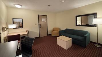 A hotel room with a desk, a chair, a sofa, a small table, a lamp, a mirror, and a door.