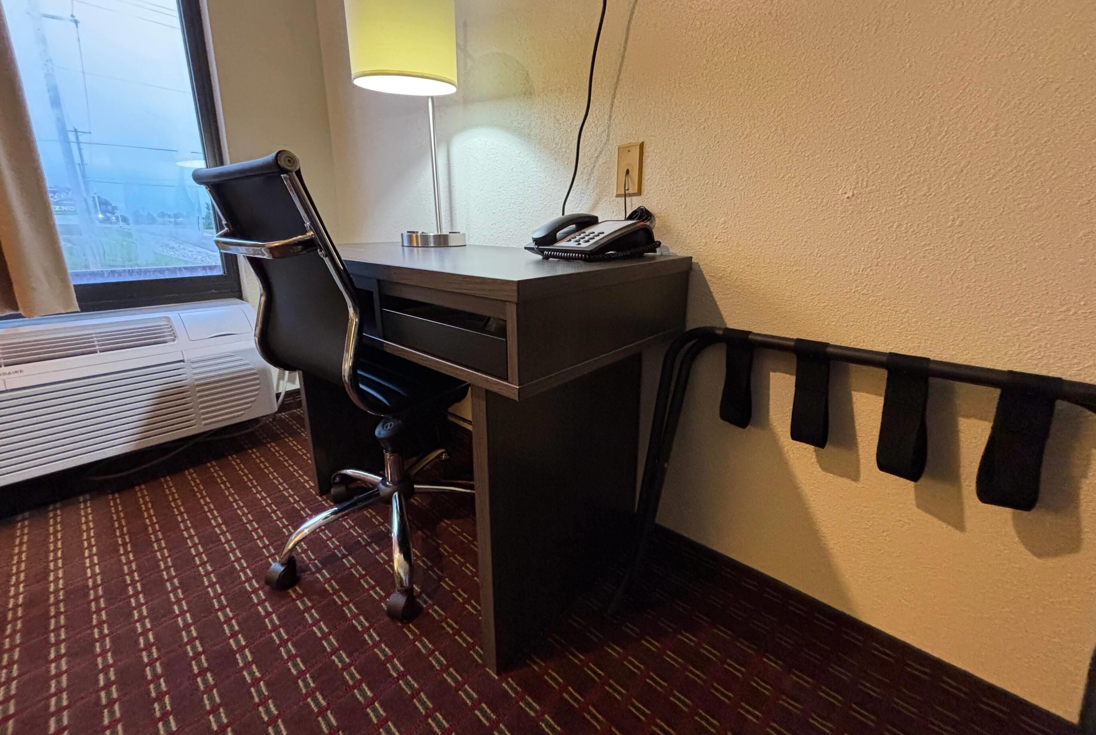 A hotel room with a desk, chair, lamp, phone, and a radiator.
