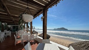 An outdoor seating area with white tables and chairs, overlooking a beach with waves and a distant island.