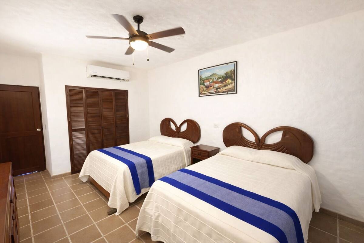 Una habitación con dos camas, ventilador de techo, equipo de aire acondicionado, armario de madera y un cuadro en la pared.