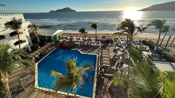 A pool area with a view of the ocean and a beach, surrounded by palm trees and lounge chairs.