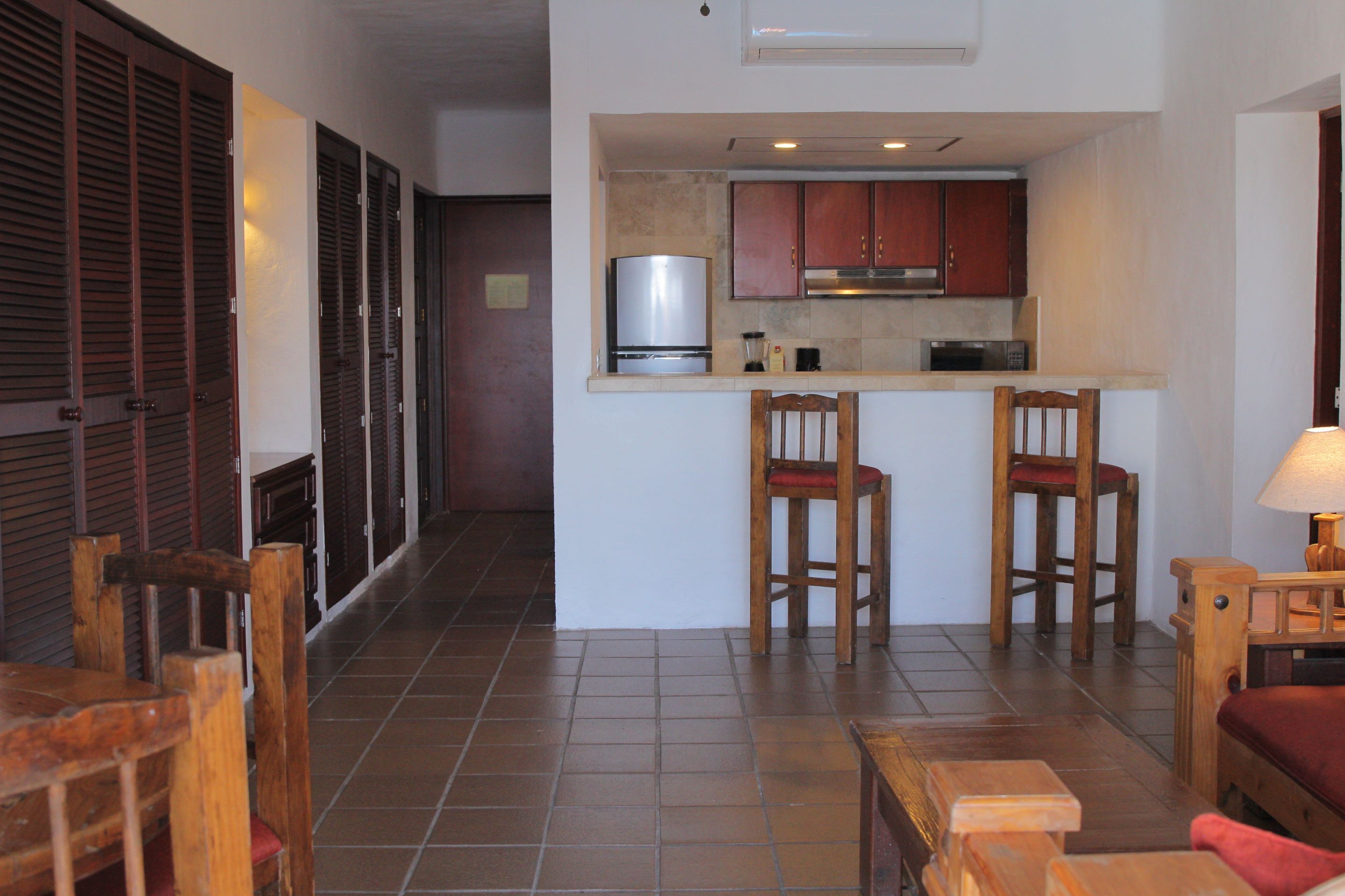 A compact kitchen with a refrigerator, microwave, and wooden cabinets. There are two wooden chairs and a small table with a lamp. The room has tiled flooring and a wooden bench.