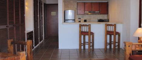 A compact kitchen with a refrigerator, microwave, and wooden cabinets. There are two wooden chairs and a small table with a lamp. The room has tiled flooring and a wooden bench.