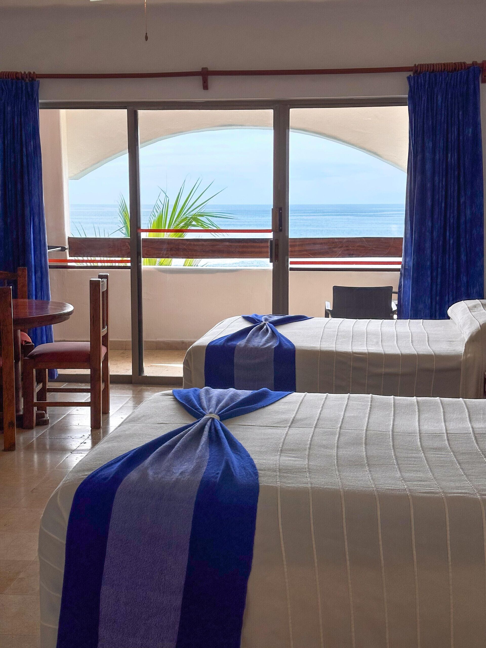 A hotel room with two beds, a wooden chair, a table, and a view of the ocean.