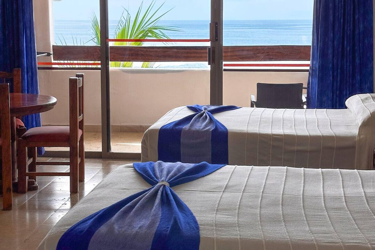 Habitación de hotel con dos camas, una silla de madera, una mesa y vista al océano.