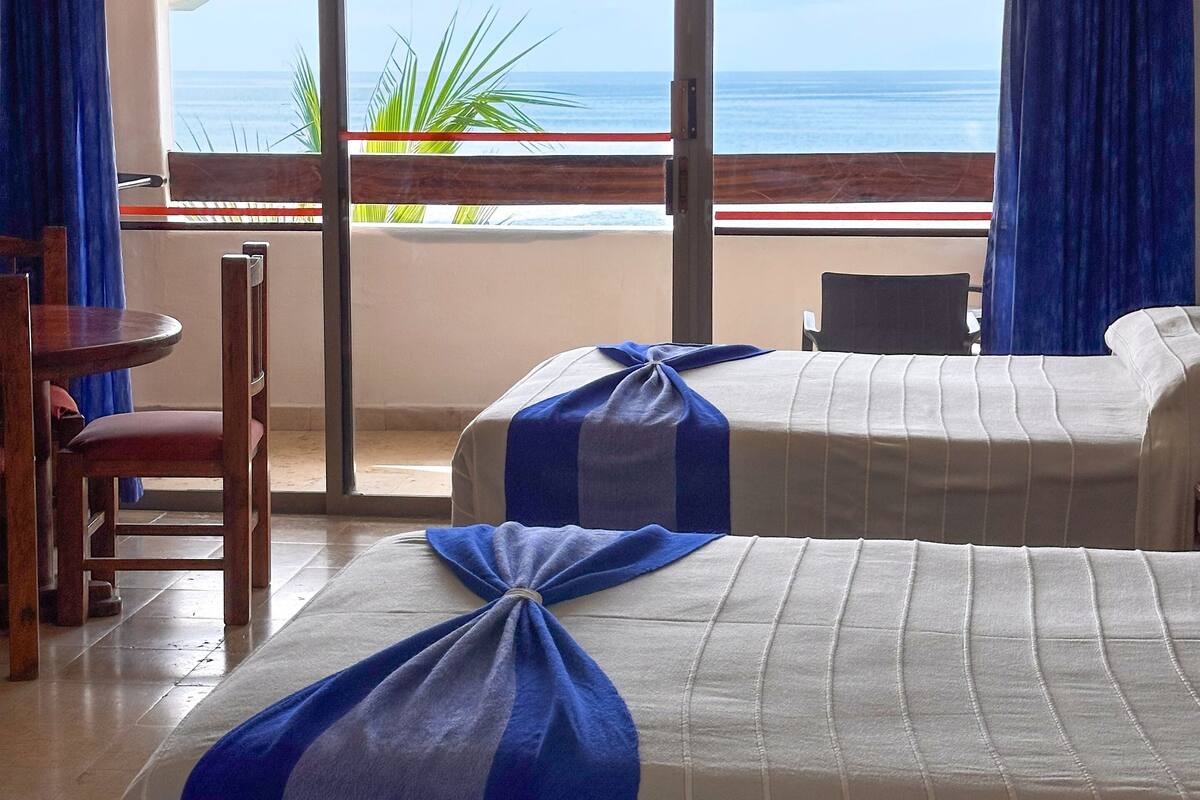 Habitación de hotel con dos camas, una silla de madera, una mesita pequeña y vistas al océano a través de un ventanal grande.
