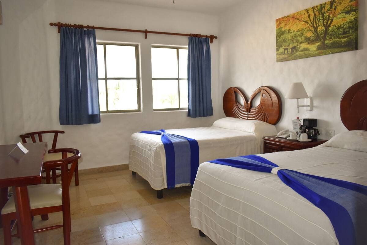 Habitación de hotel con dos camas, cabecera de madera, escritorio, sillas, ventana con cortinas, un cuadro en la pared y una mesita de noche con lámpara y teléfono.