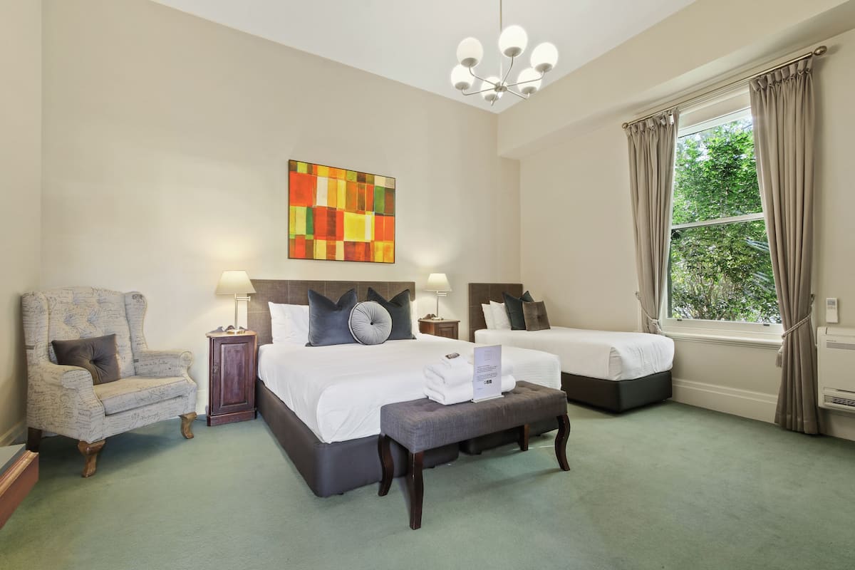 Une chambre d’hôtel avec deux lits, un grand fauteuil, un bureau et une fenêtre avec des rideaux.