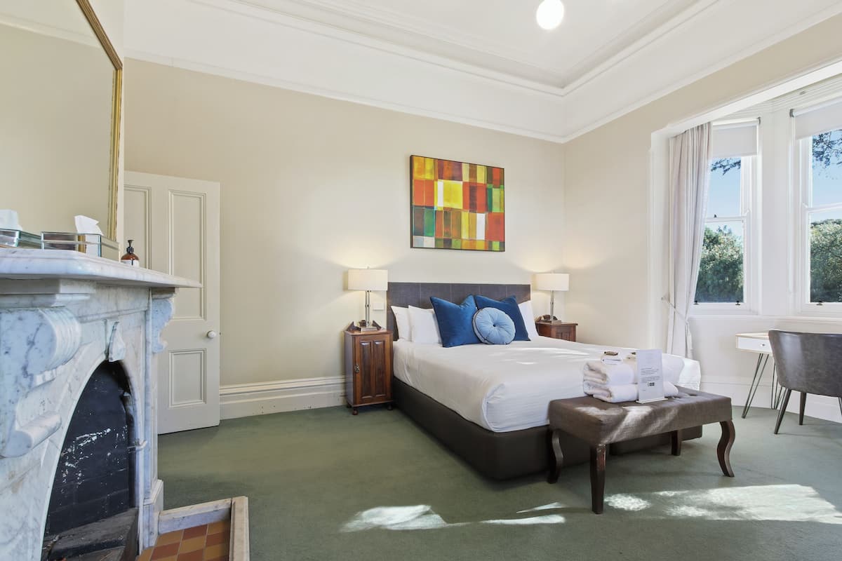 Une chambre d’hôtel avec un lit, des lampes de chevet, une cheminée, un bureau et une fenêtre donnant sur un beau paysage.