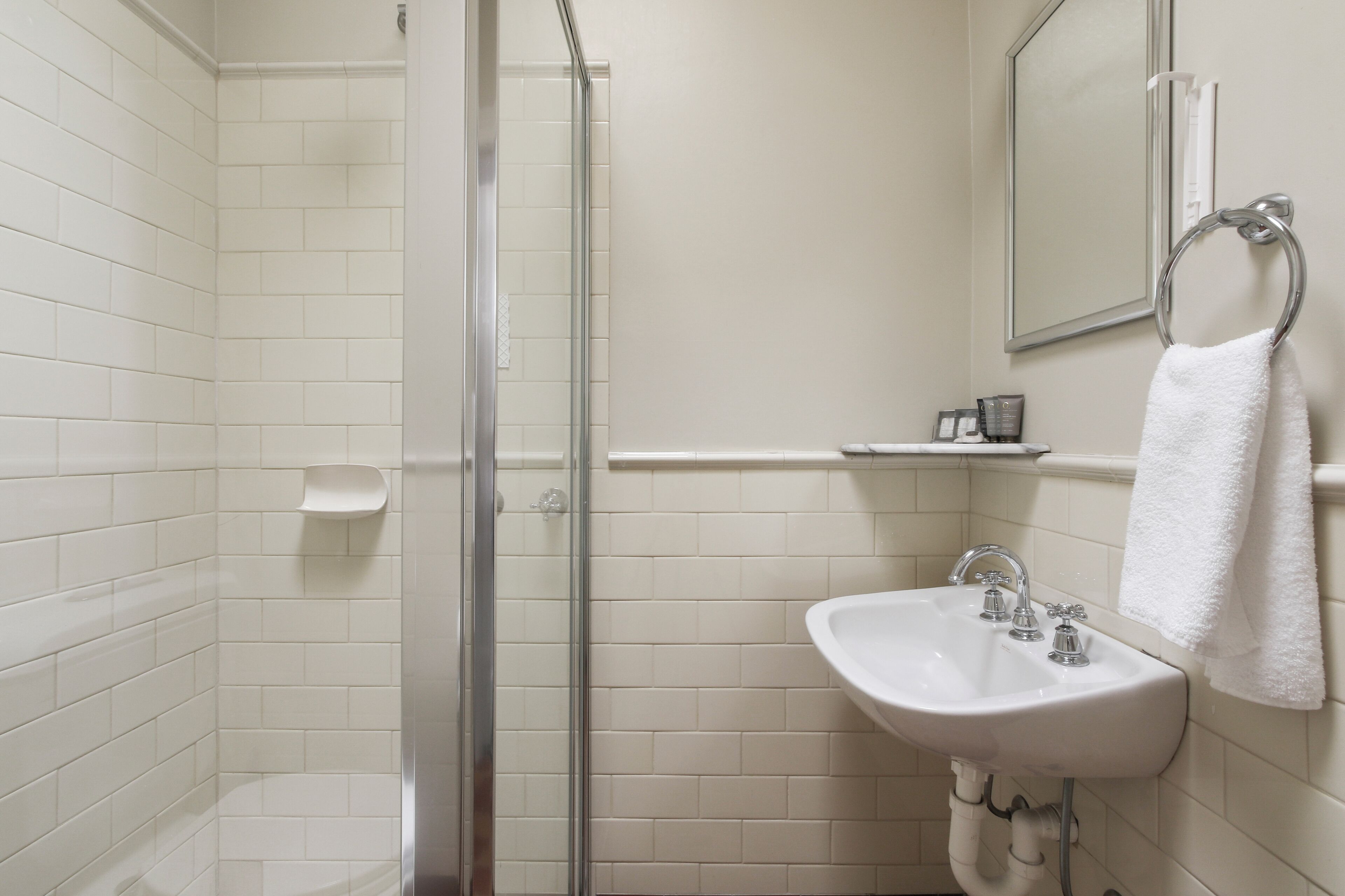 Un baño con paredes de azulejos blancos, mampara de ducha de vidrio, lavamanos blanco con dos grifos y un perchero para toallas en la pared.