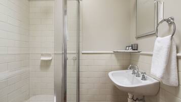 Un baño con paredes de azulejos blancos, mampara de ducha de vidrio, lavamanos blanco con dos grifos y un perchero para toallas en la pared.