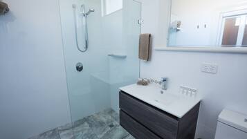 Un baño moderno con ducha envuelta en vidrio, un lavabo blanco con tocador de madera oscura y un espejo sobre el lavabo.