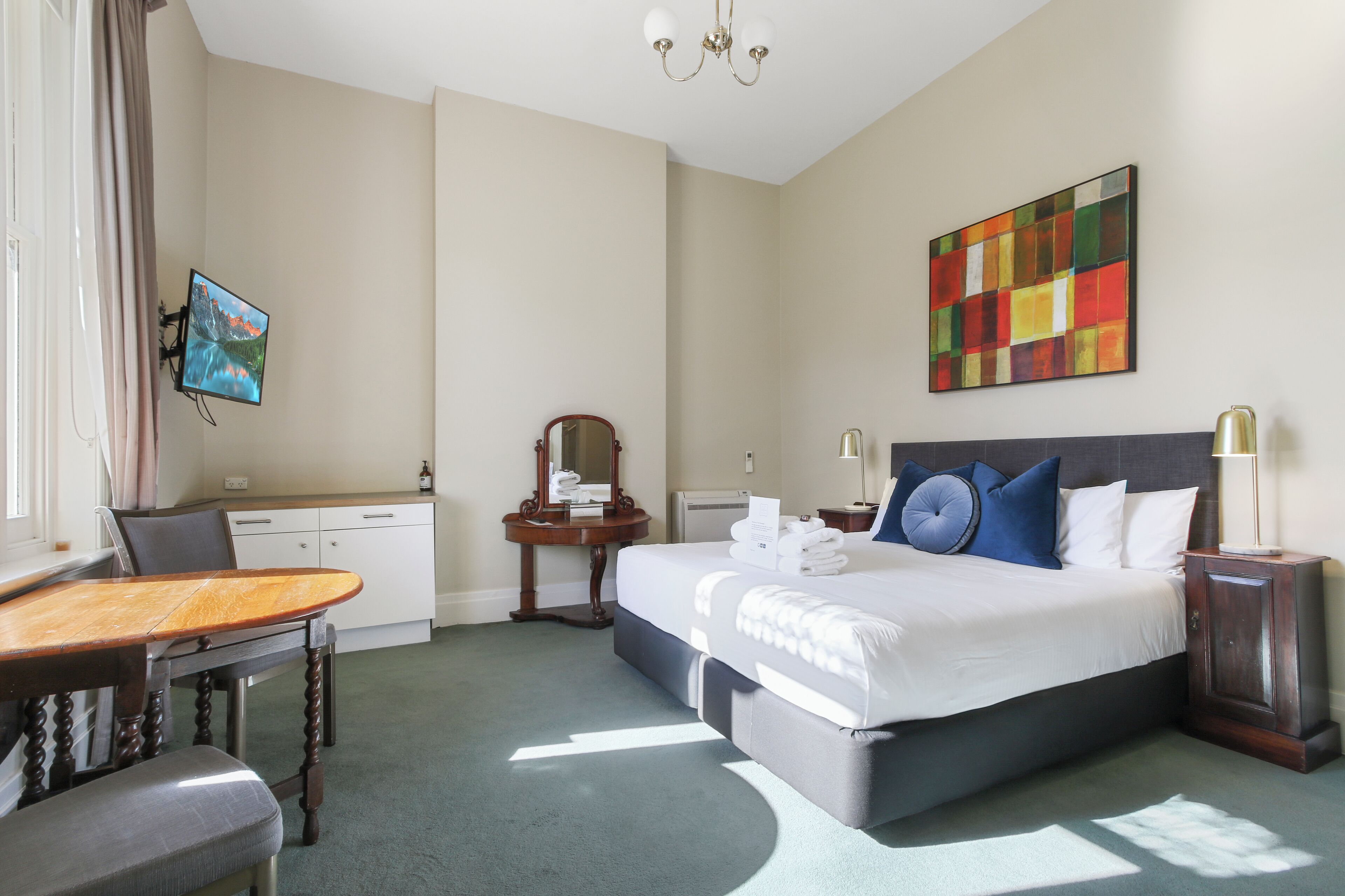 A hotel room with a bed, a desk, a chair, a TV, and a painting on the wall.