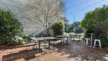 An outdoor patio with brick flooring, metal tables and chairs, string lights, and greenery.