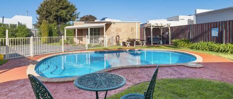 A swimming pool with a patio area, including a table and chairs, surrounded by a fence and greenery.