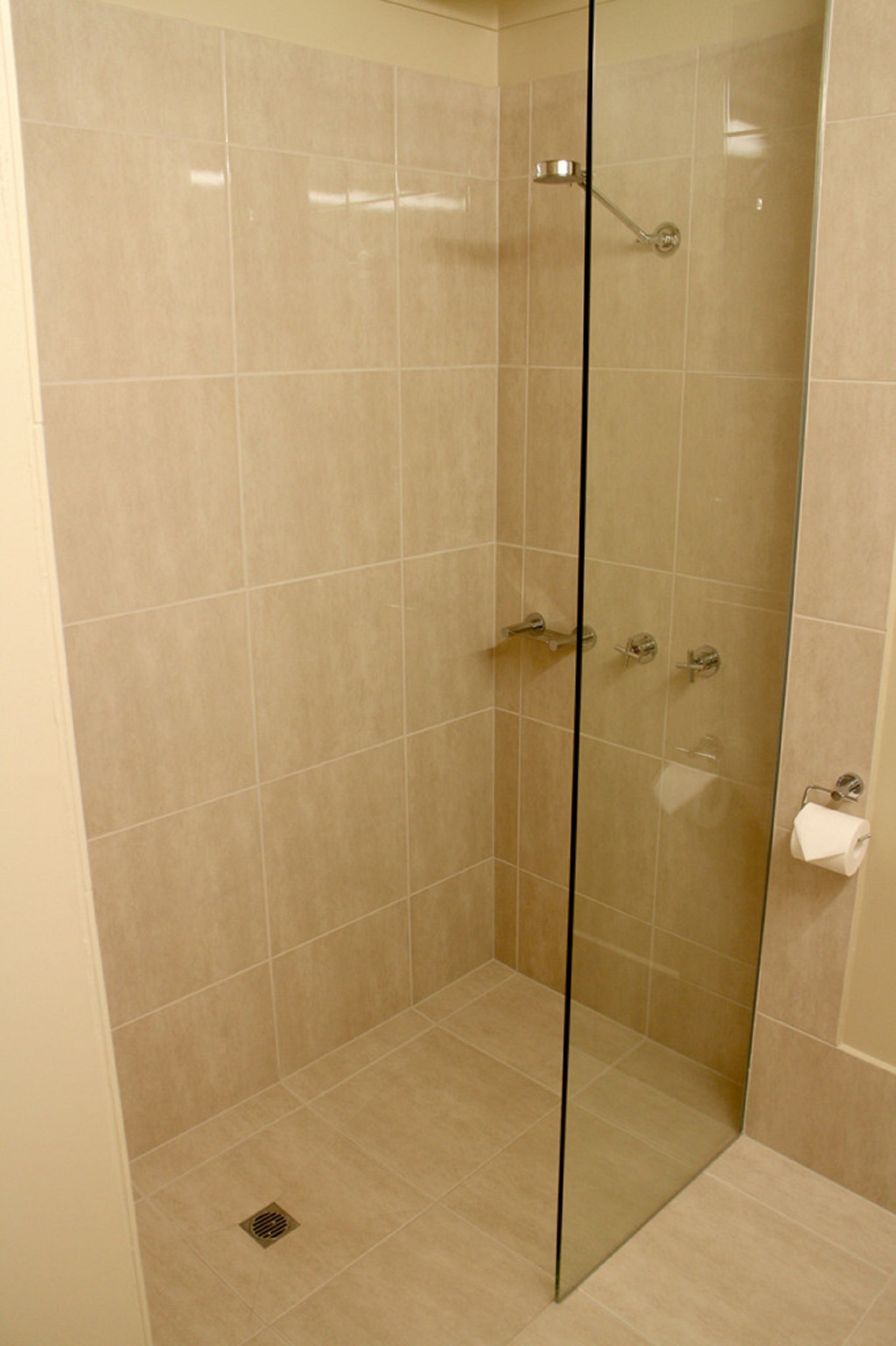 A glass-enclosed shower stall with beige tiles, a handheld showerhead, and a toilet paper holder.