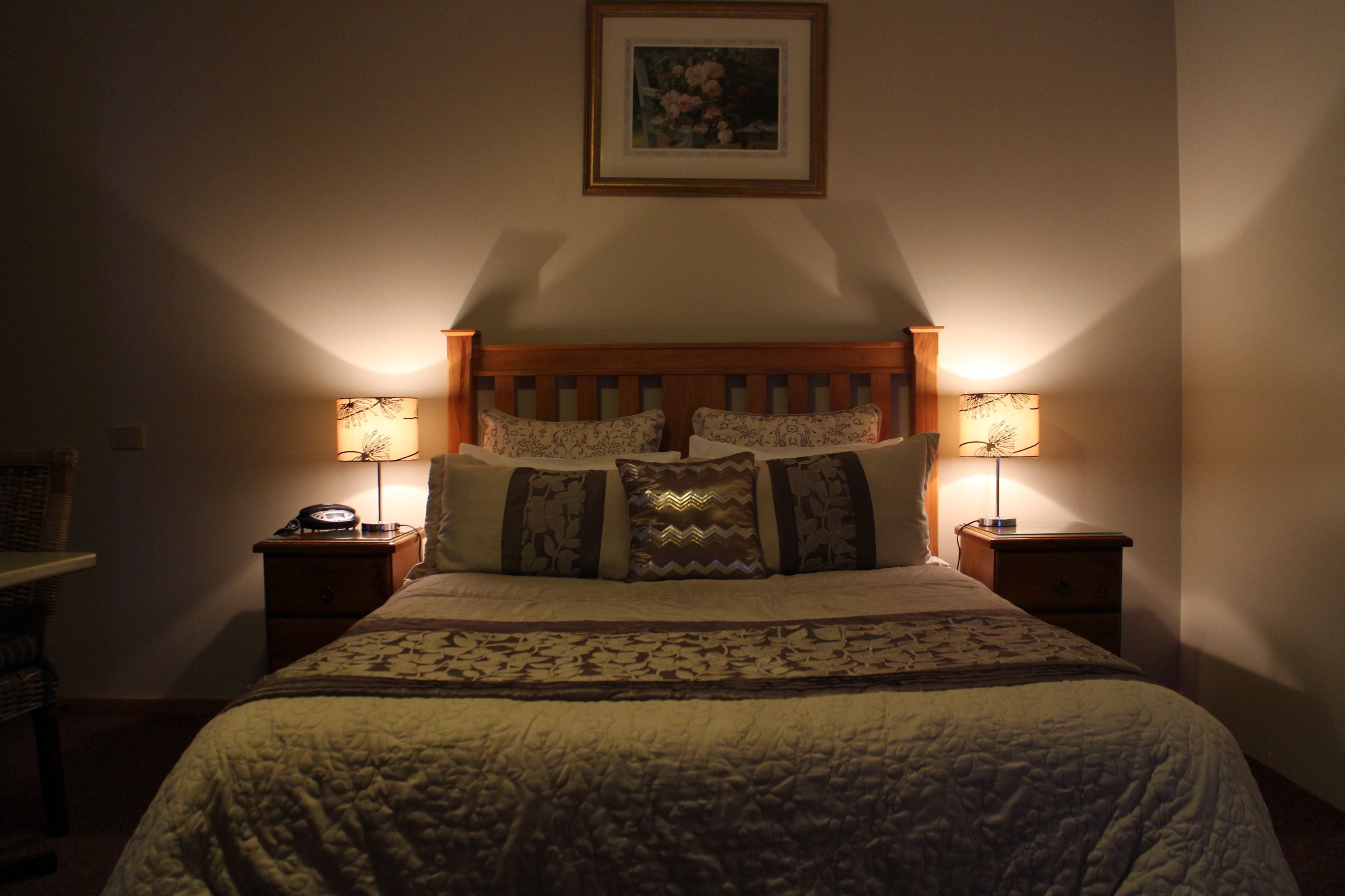 Een net opgemaakt bed met een bedsprei en kussens, aan weerszijden geflankeerd door nachtkastjes met lampen, in een kamer met een ingelijst beeld op de muur.
