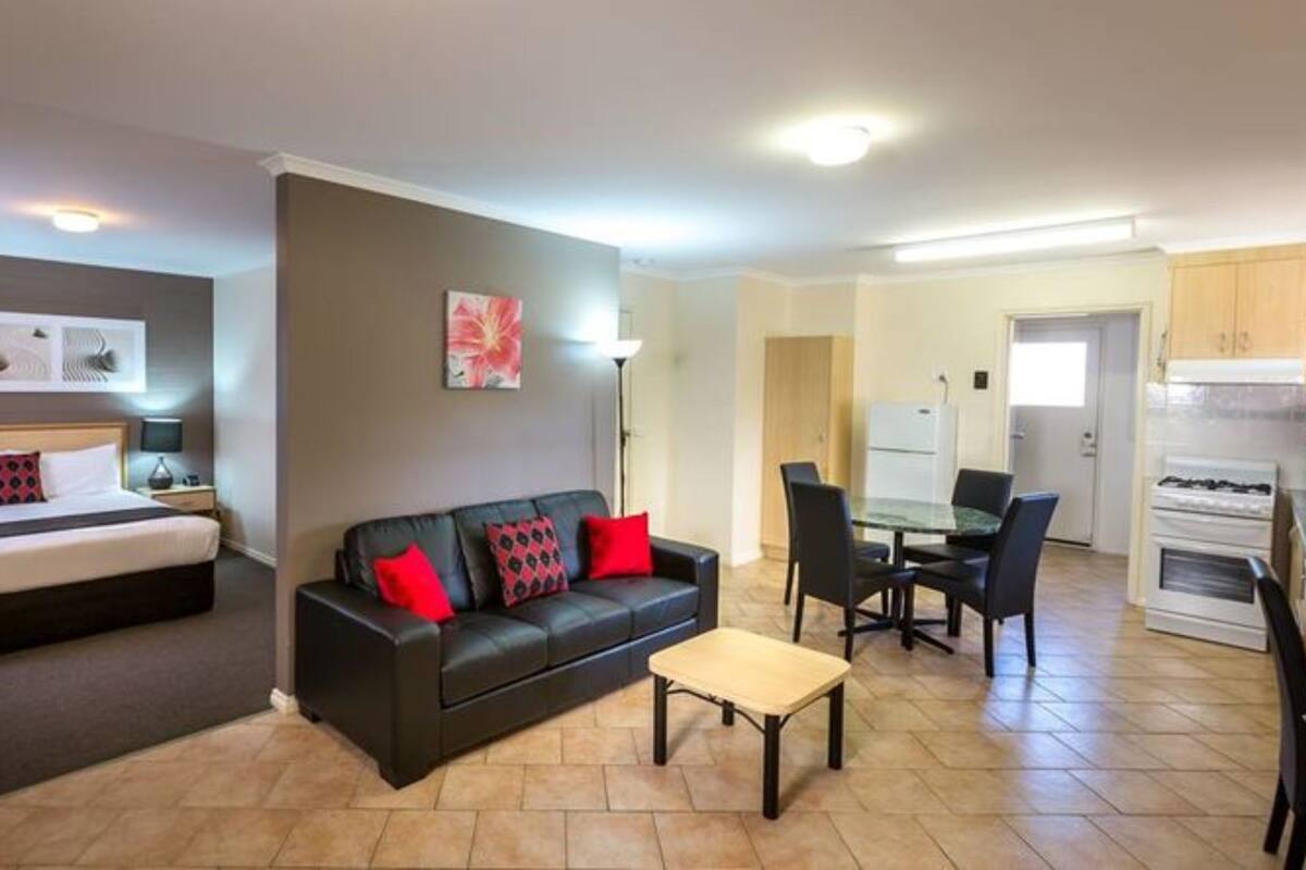 A hotel room with a bed, a sofa, a dining table with chairs, a kitchen area, and a hallway.