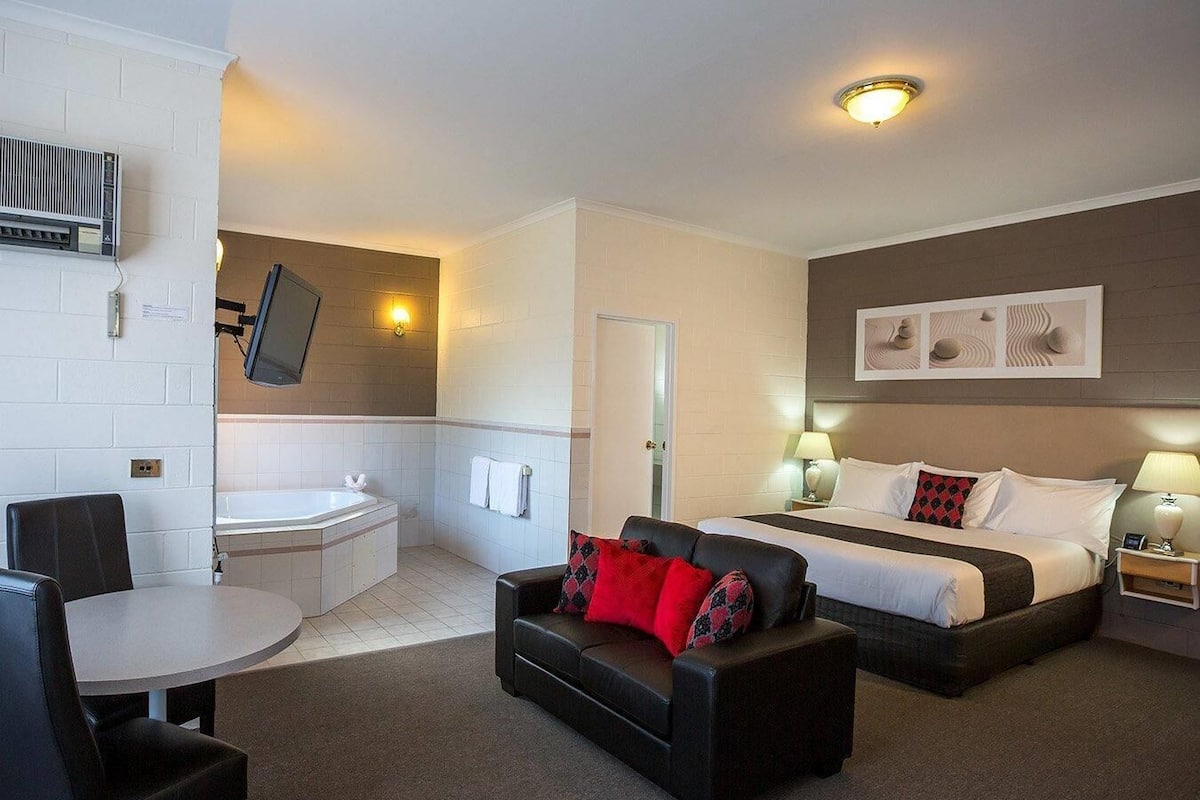 Ein Hotelzimmer mit Bett, Sofa, kleinem Tisch, Fernseher und einem Badezimmer. An der Wand ist ein Flachbildfernseher angebracht.