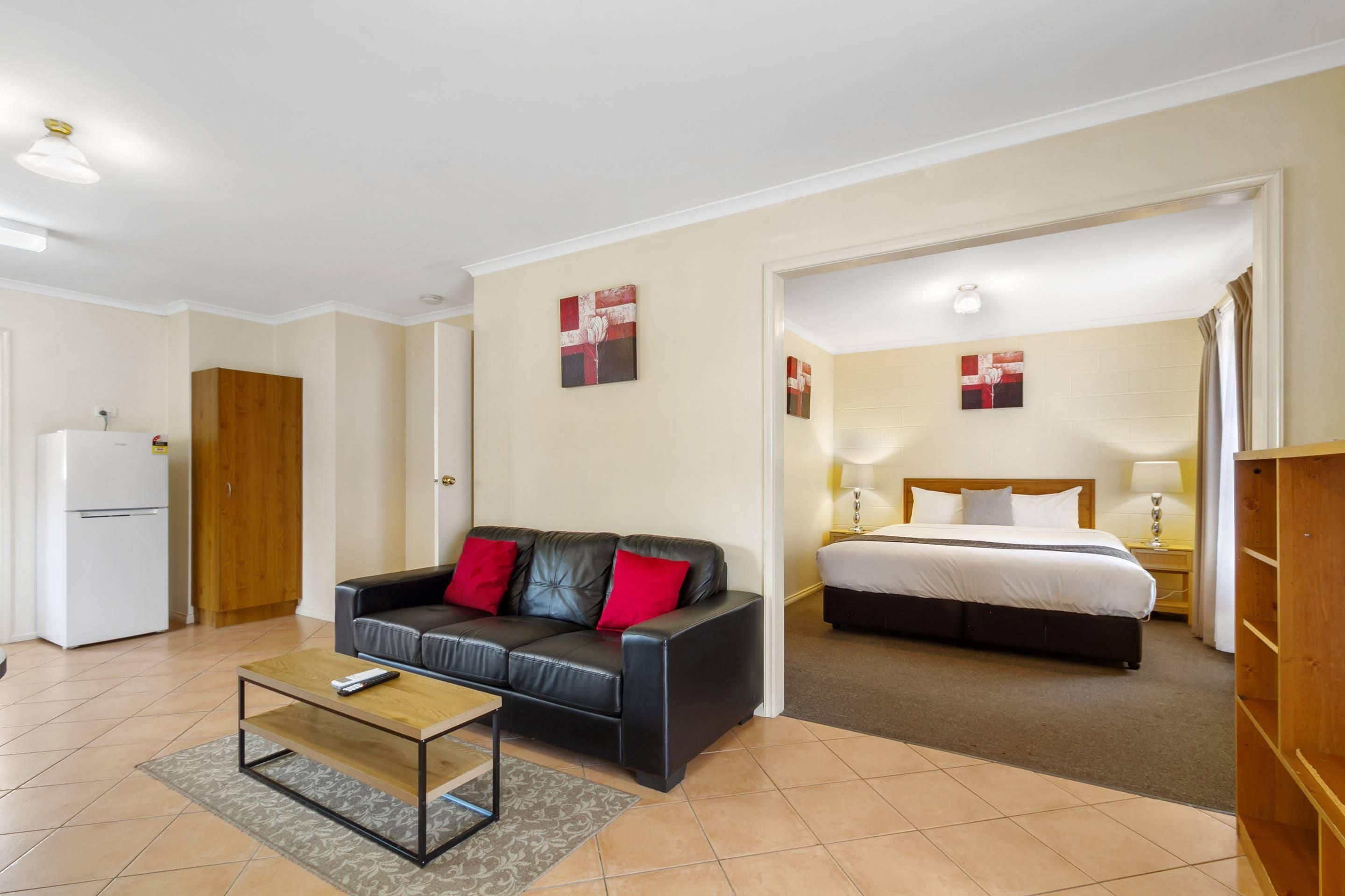 A hotel room with a black leather sofa, a wooden coffee table, a bed with white bedding, and a small bedside table with a lamp.