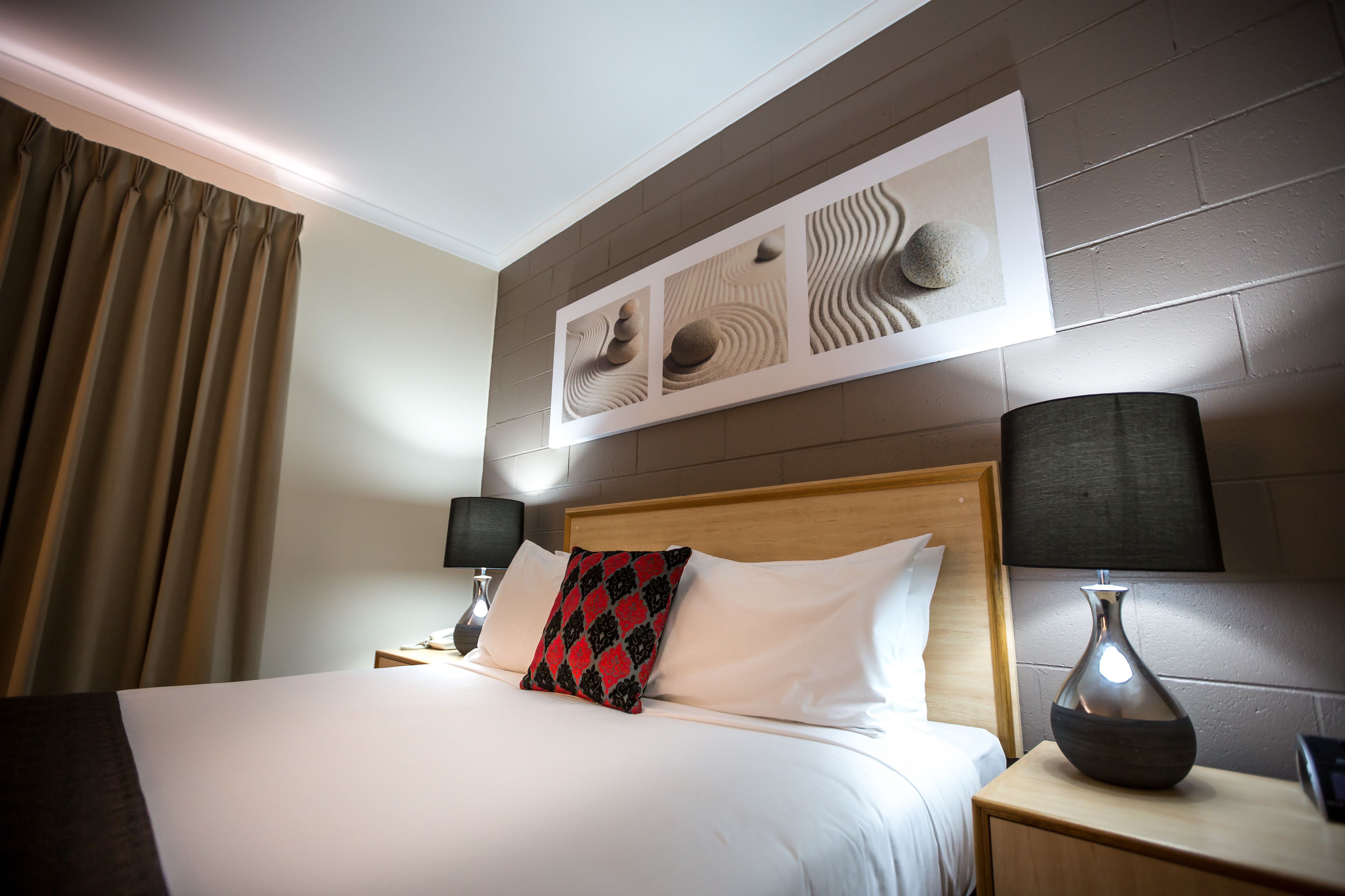 Une chambre d’hôtel comprenant un lit, deux tables de chevet, une commode, une lampe, un tableau accroché au mur et un rideau.