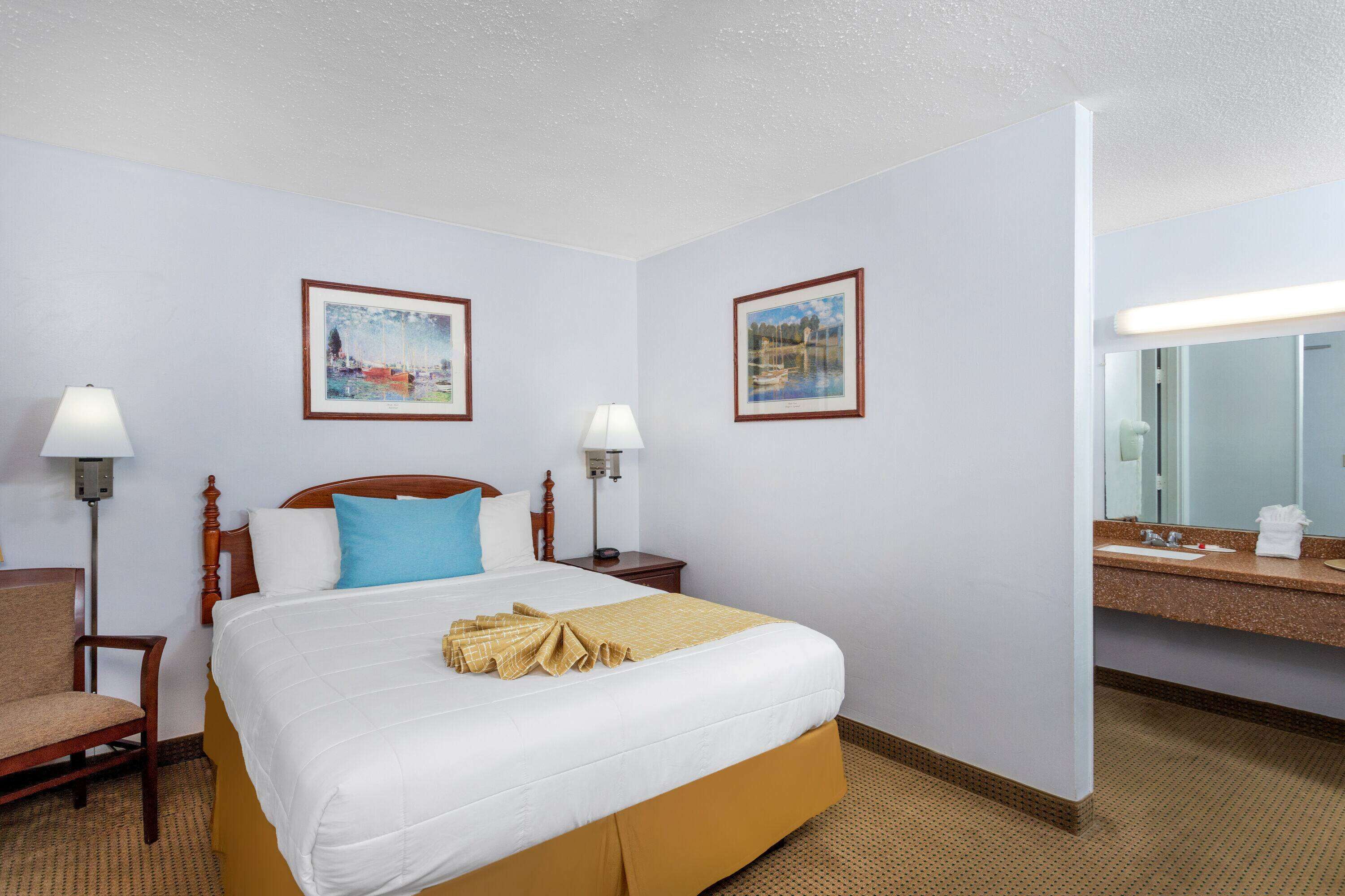 Habitación de hotel con una cama grande, dos mesitas de noche con lámparas, una silla de madera, un baño con lavamanos y espejo, y dos cuadros enmarcados en las paredes.