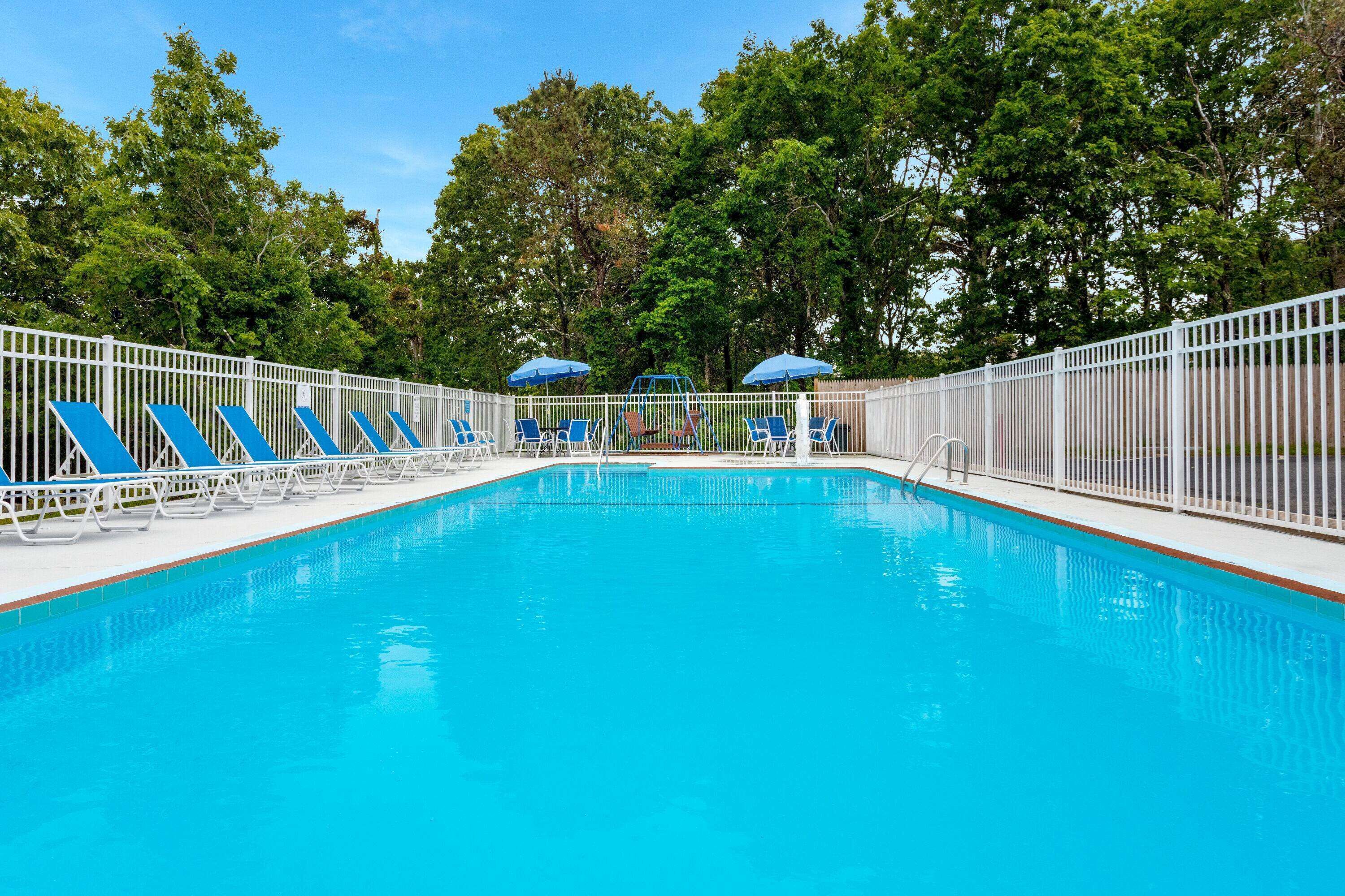 Una piscina con sillas y sombrillas azules, rodeada por una valla blanca y árboles.