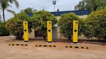 Three yellow RAA electric vehicle charging stations with a building and trees in the background.