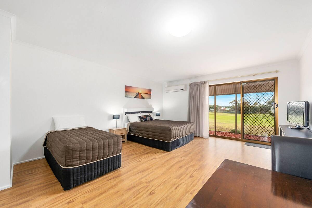 A hotel room with two beds, a desk, a TV, and a large window with a view of a green field.