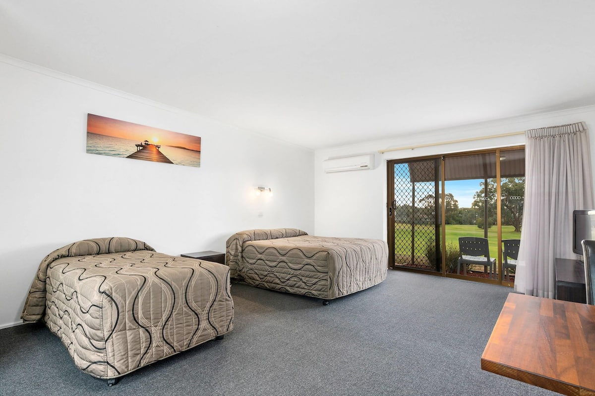 A hotel room with two beds, a painting of a sunset over water, a sliding glass door to a balcony, and a wooden table.